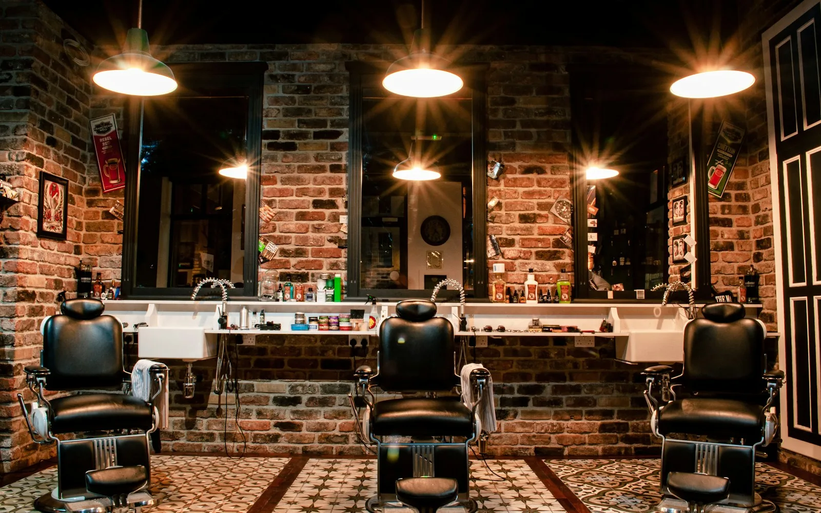 Trzy czarne skórzane fotele barberskie przed lustrami na tle ceglanej ściany