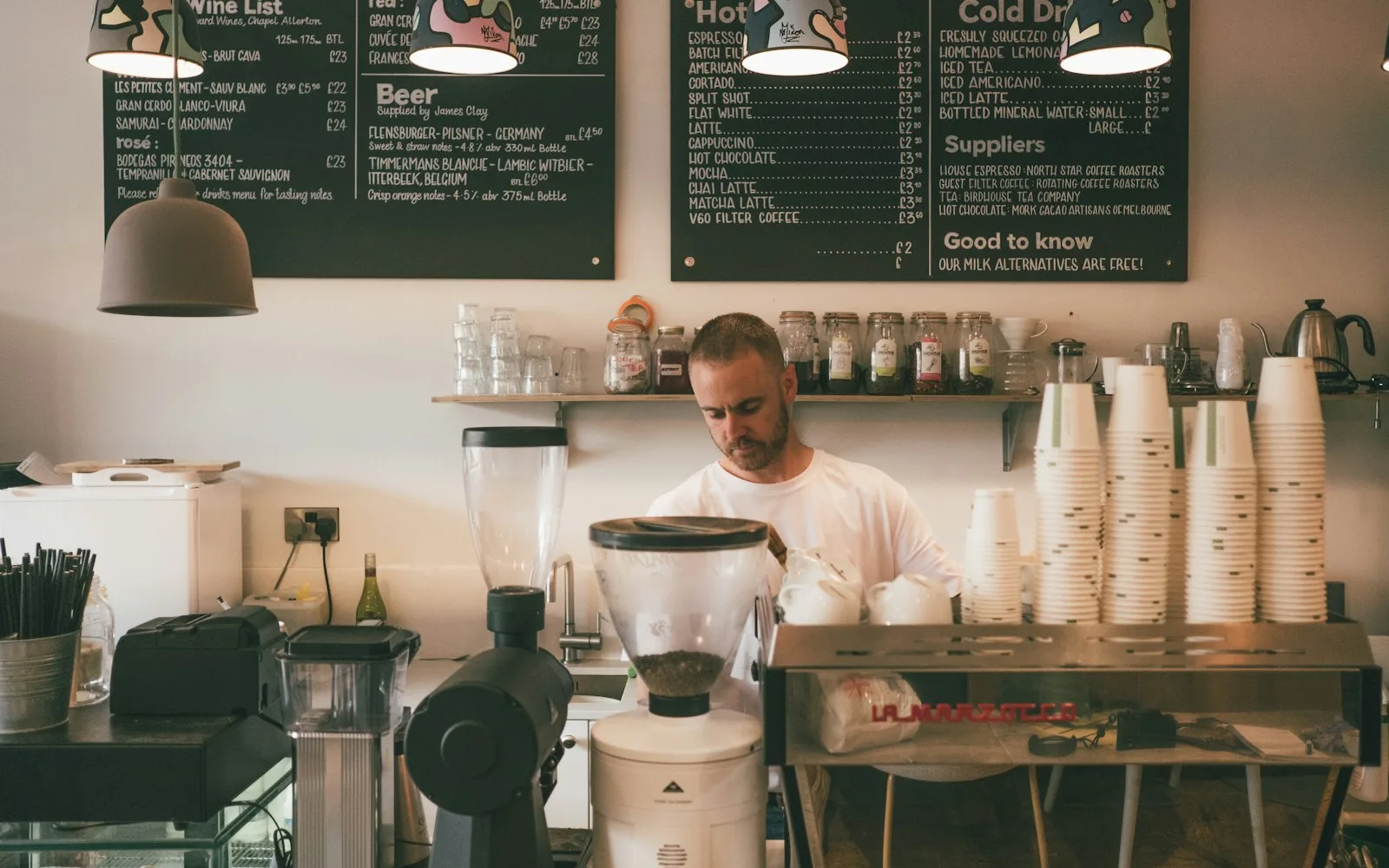 Właściciel kawiarni za ladą z ekspresem, kubkami i tablicą z menu